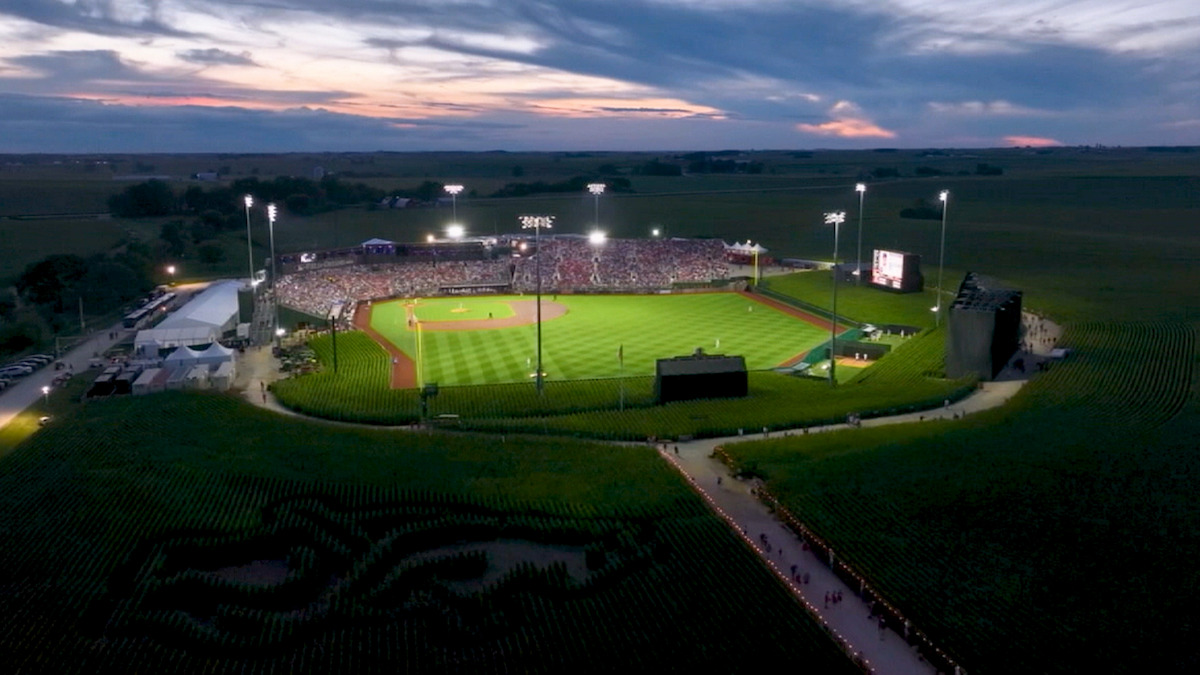 Aerial view of a baseball stadium surrounded by cornfields at sunset, stadium lights on, crowd in stands, rural setting, dramatic sky, paths through the fields, and large design carved into the corn near the entrance.