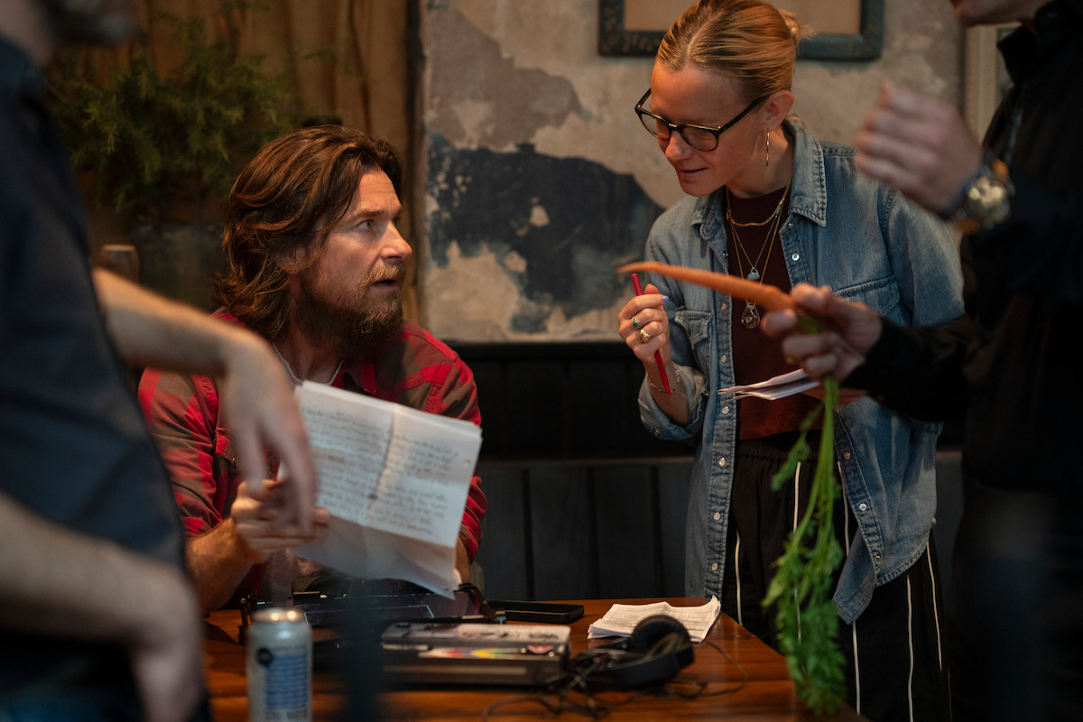 Man with long hair in plaid shirt and woman in glasses discuss papers in a rustic indoor setting; hands with a carrot and papers are visible, with a can and other items on a wooden table, suggesting a collaborative meeting.