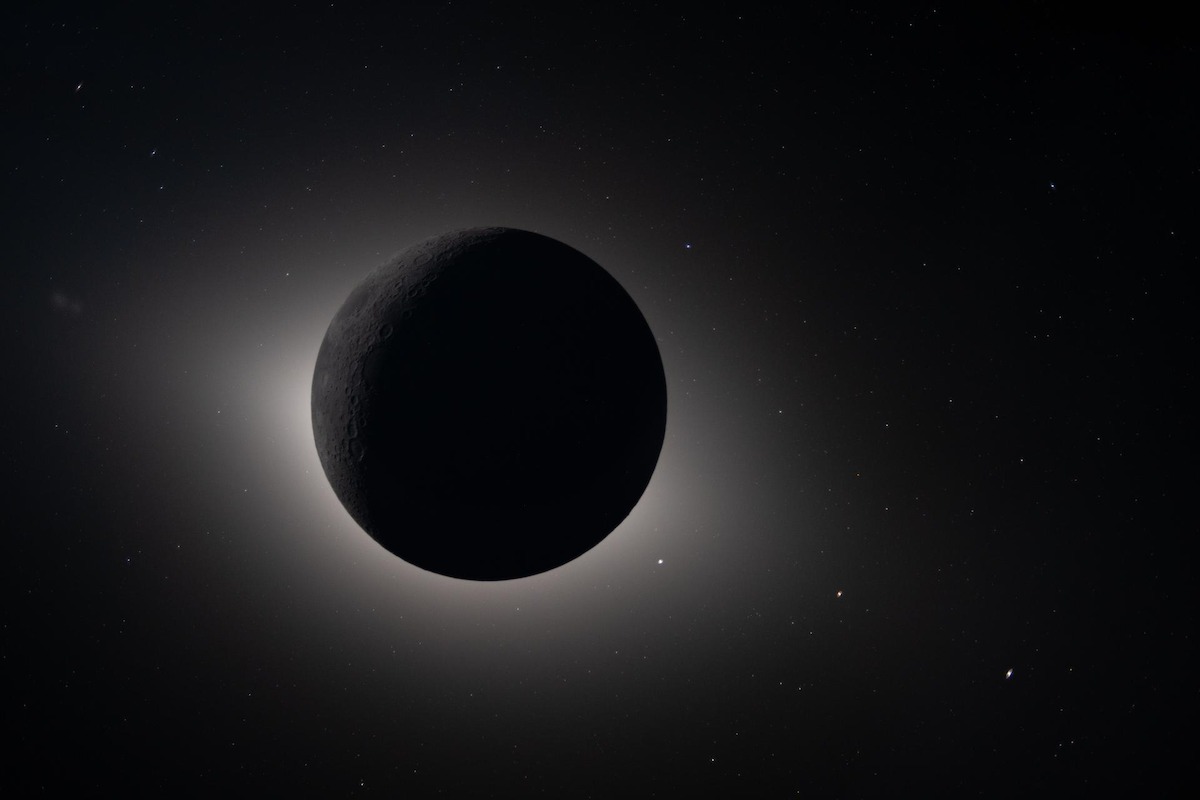 A dark celestial body, likely the moon, is seen during a total solar eclipse with the sun's corona glowing around it against a backdrop of stars in space.