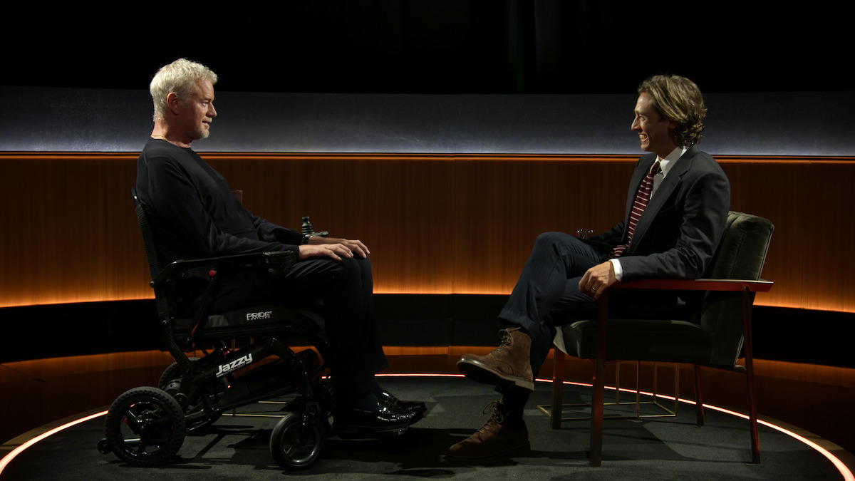 Eric Dane and Brad Falchuk sit facing each other in a dimly lit studio, one in a wheelchair and the other in a chair, engaged in conversation on a stage with warm, modern lighting and a minimalist background.