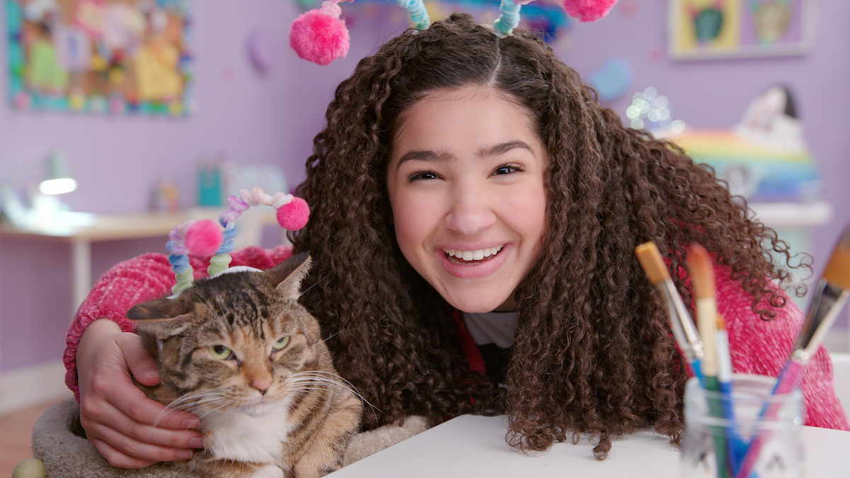 Smiling girl with curly hair and cat wearing matching headbands in a colorful, playful room with art supplies and rainbow decor, suggesting a fun and creative environment.