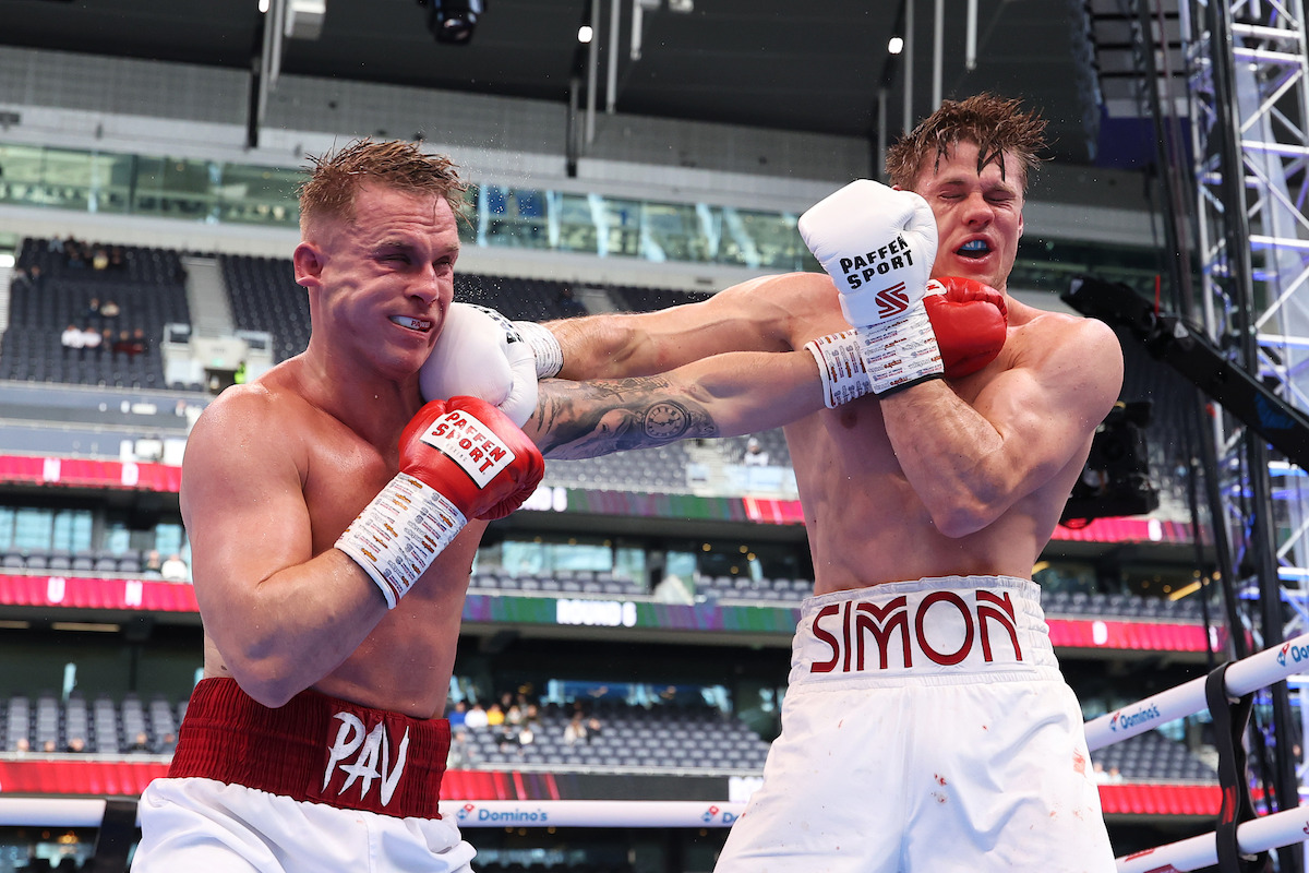 Two male boxers in a boxing ring during a match, one landing a punch to the other's face. The setting is a large indoor arena with spectators in the background.