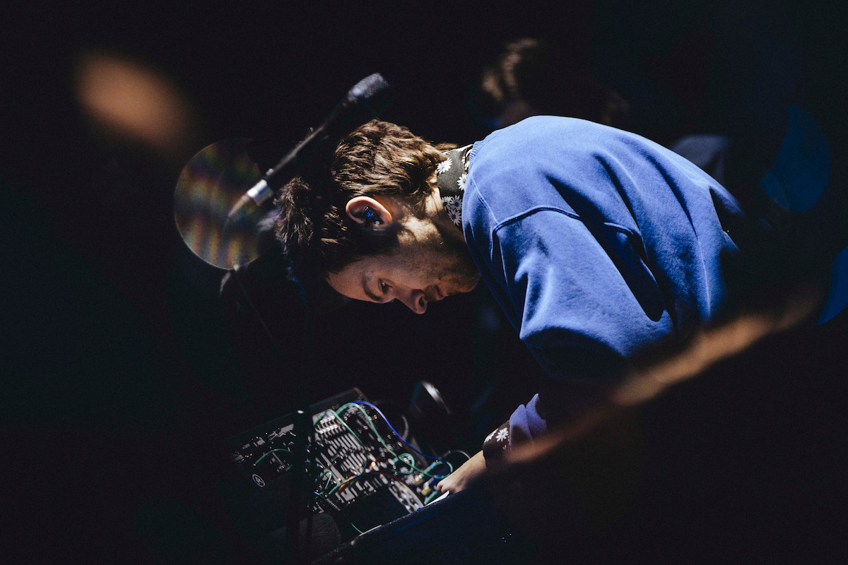 Harry Styles in a blue sweatshirt focused on adjusting audio equipment in a dimly lit environment, possibly a music performance or studio setting.