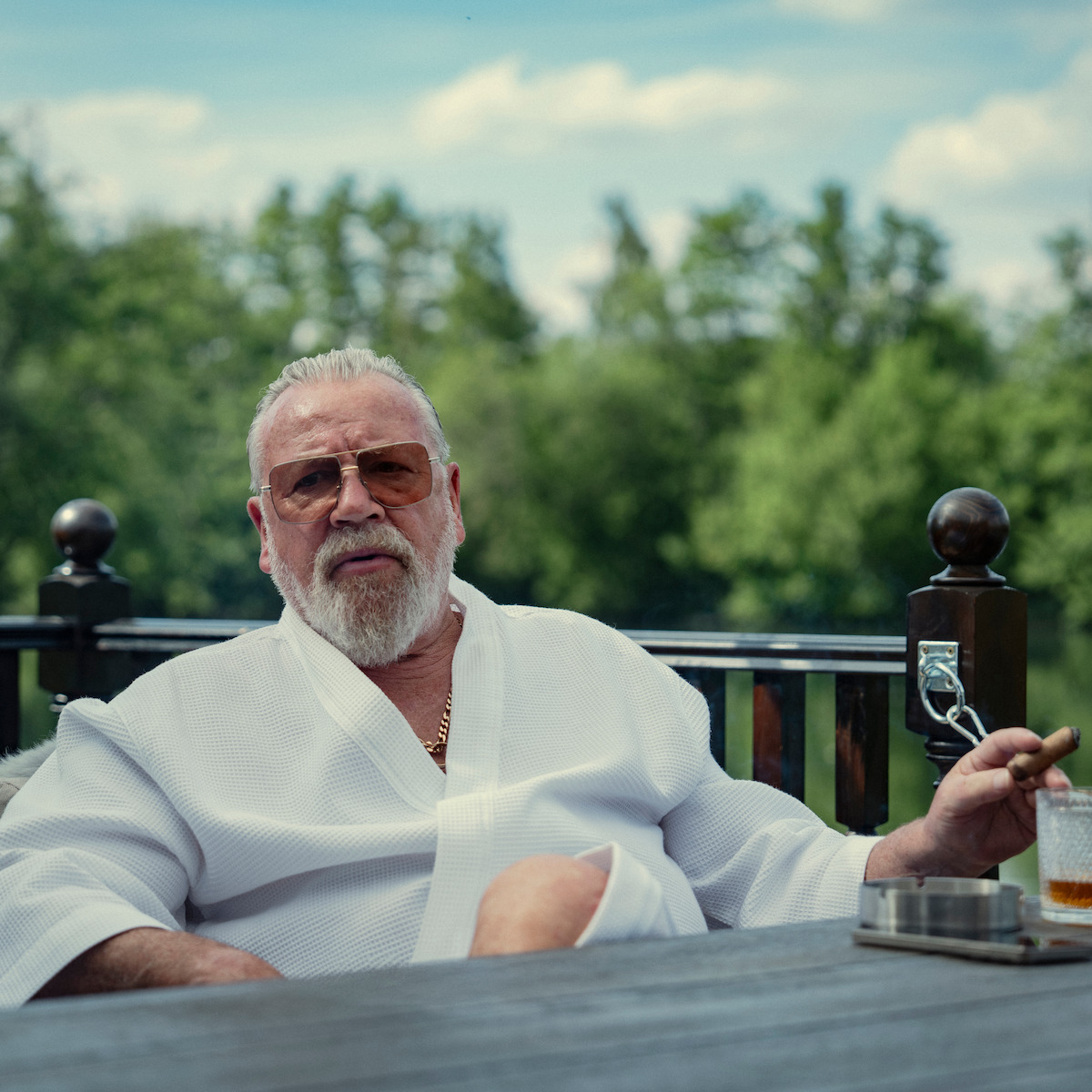 Older man with white beard in a robe, wearing sunglasses, sits outdoors on a patio with trees in the background, holding a cigar and a drink, relaxing at a table on a sunny day.