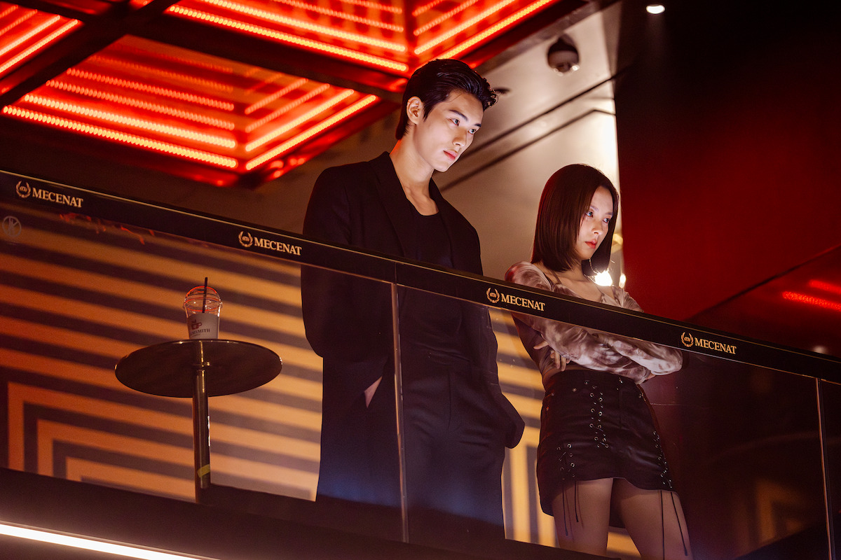 A stylish man and woman stand on a modern balcony at a dimly lit nightclub with red neon lights, leaning on a glass railing near a table with a drink, looking thoughtful or serious.