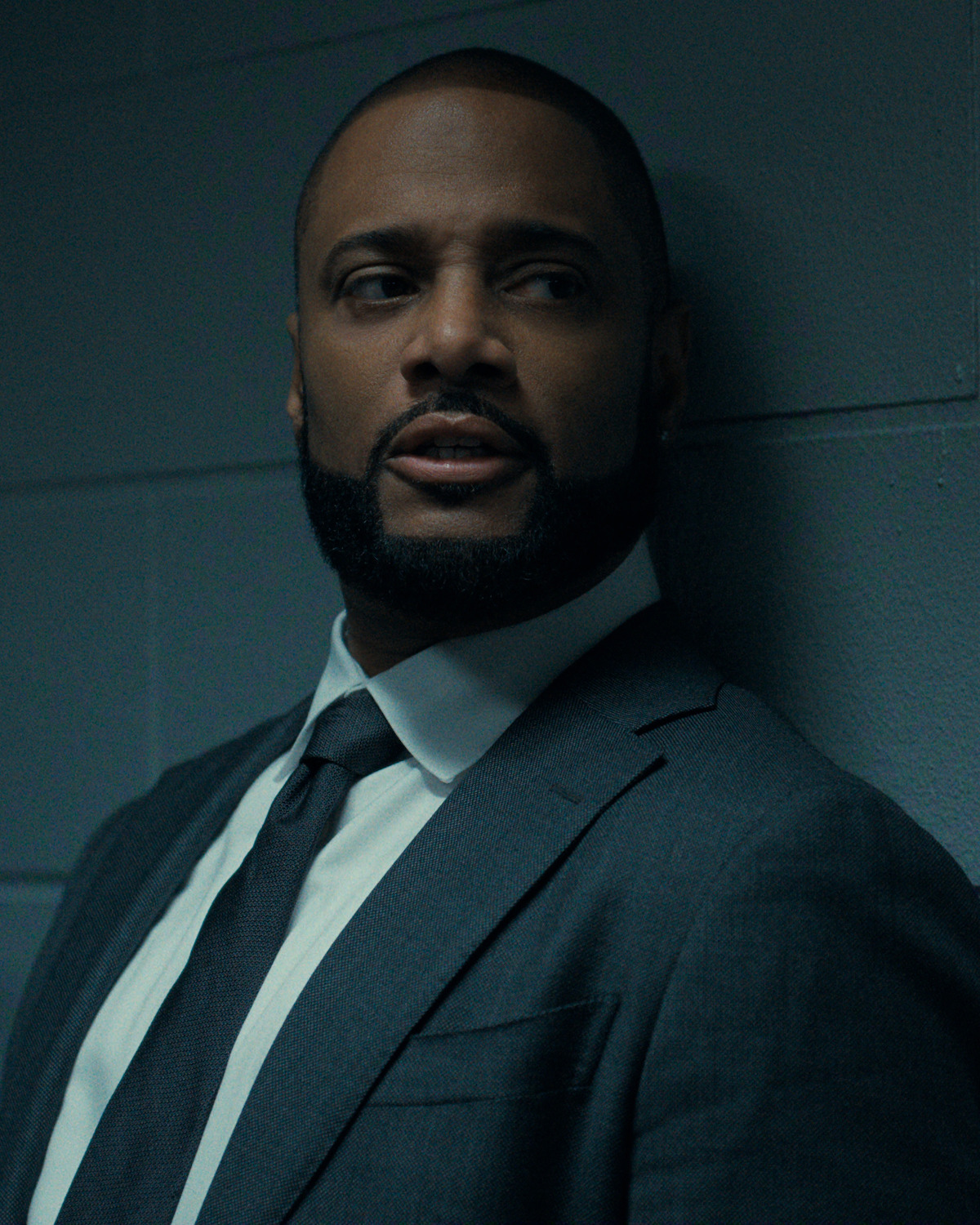 Man in a suit and tie leaning against a dimly lit, gray cinder block wall, looking to the side with a serious expression, suggesting a tense or dramatic moment in an indoor setting.