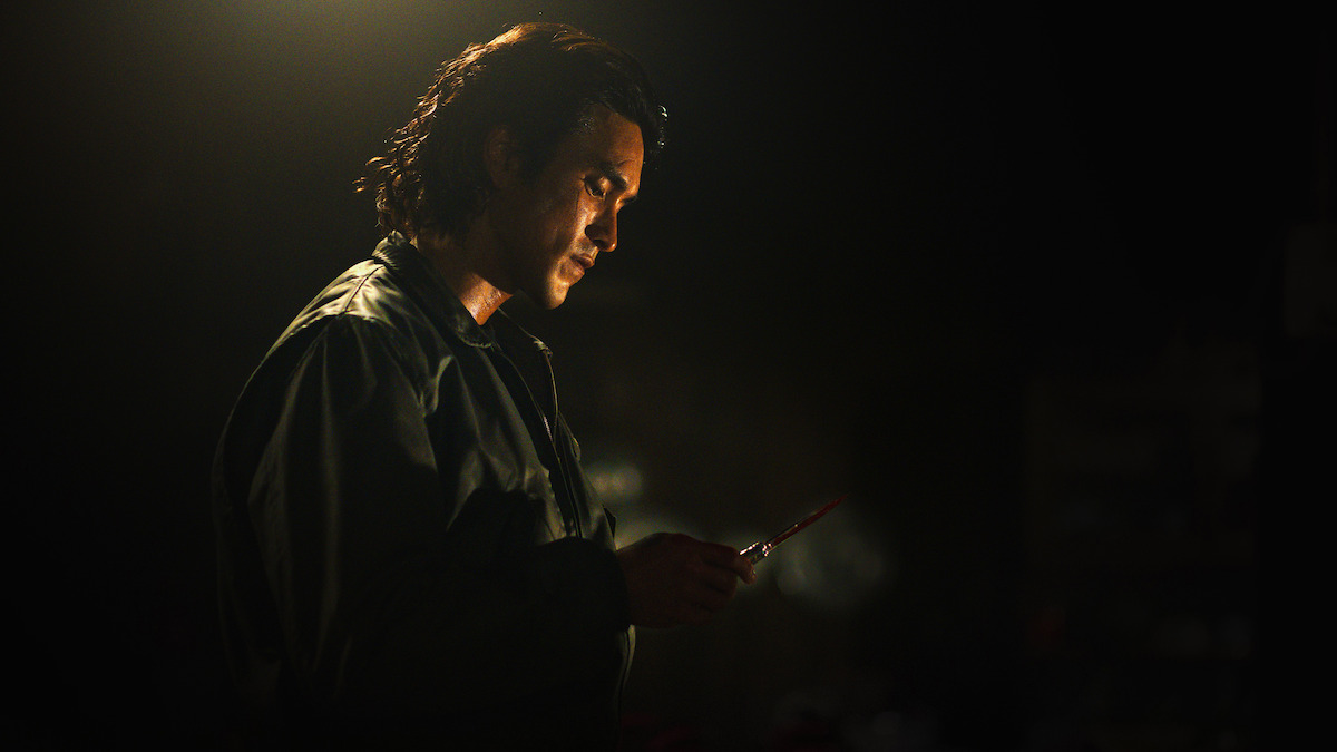 A man stands in a dimly lit, shadowy room holding a knife, illuminated dramatically from above, creating a tense and suspenseful atmosphere.