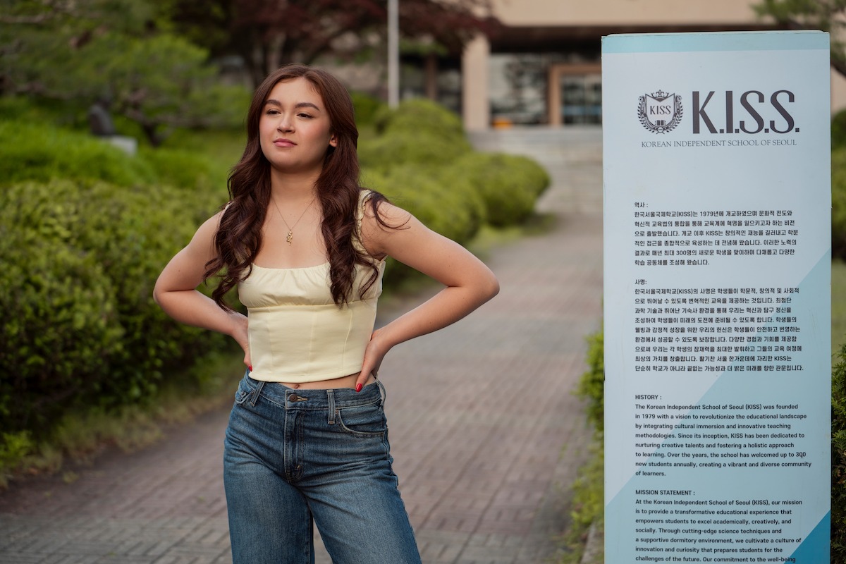 Young woman in a yellow top and jeans stands confidently outdoors on a path near green bushes and a sign with the K.I.S.S. logo and informational text, possibly on a school or campus setting.