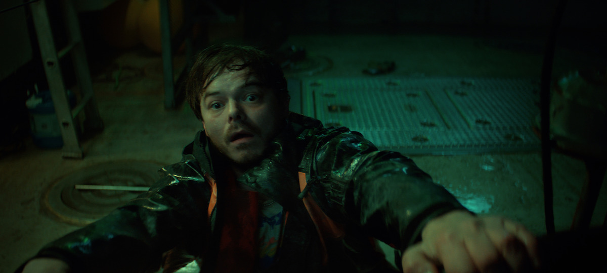 A frightened man in a raincoat clings to a metal railing inside a dimly lit, industrial room with wet floors and scattered tools, suggesting tension or danger in the environment.