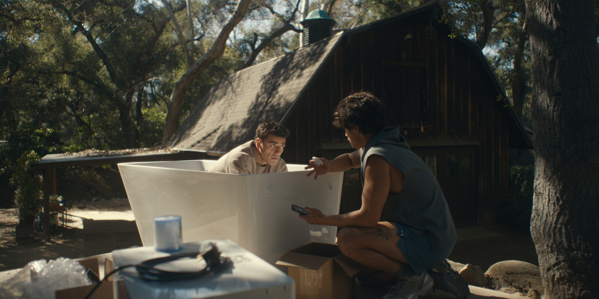 Two people assembling a large white panel outdoors near a rustic wooden cabin surrounded by trees, with tools and boxes on the ground, in a sunny forest setting.