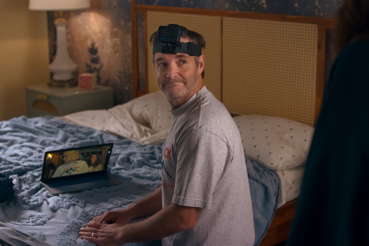 Man wearing a head-mounted camera sits on a bed in a cozy, softly lit bedroom with a tablet on the blanket, bedside table with lamp and decor in the background.