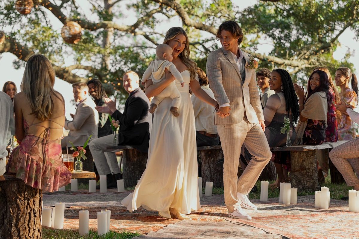 Bride and groom holding a baby walk down the aisle outdoors, surrounded by seated guests under trees, decorated with candles and natural wooden benches, during a sunny wedding ceremony.