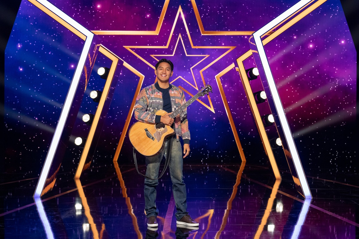 JR De Guzmán se encuentra en un escenario brillantemente iluminado sosteniendo una guitarra acústica, con un diseño en forma de estrella y un fondo colorido y estrellado, que sugiere un espectáculo de talentos o un escenario de actuación.