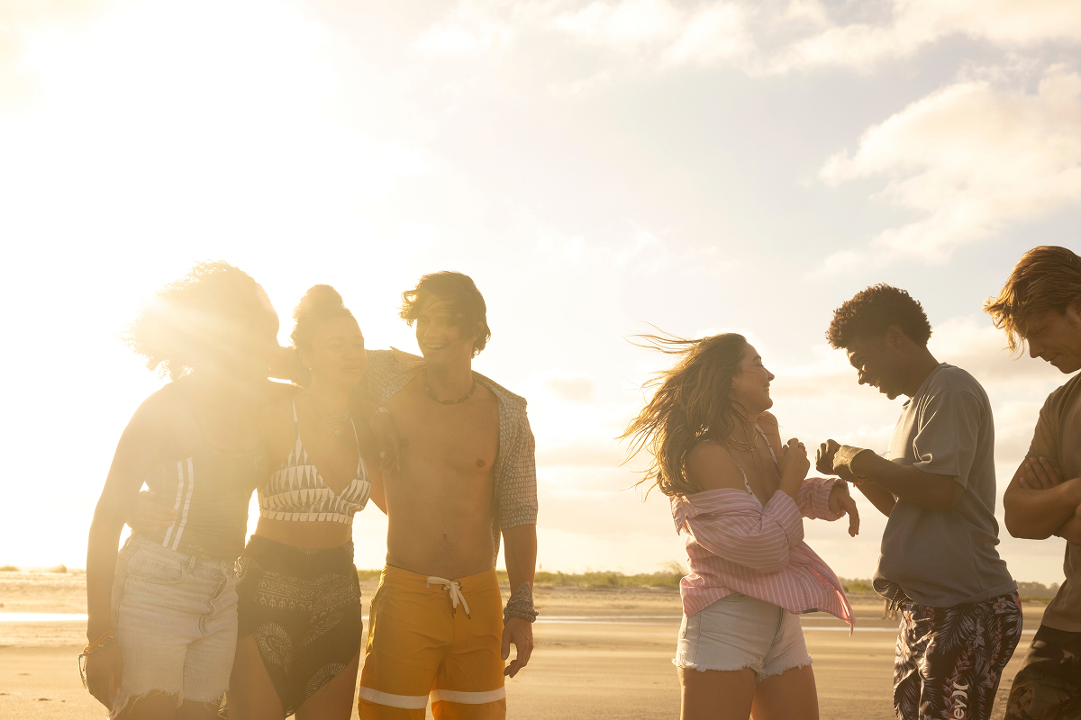 Group of young people enjoying a sunny day at the beach, laughing and talking, with sand and blue sky in the background, sunlight creating a warm, relaxed atmosphere.