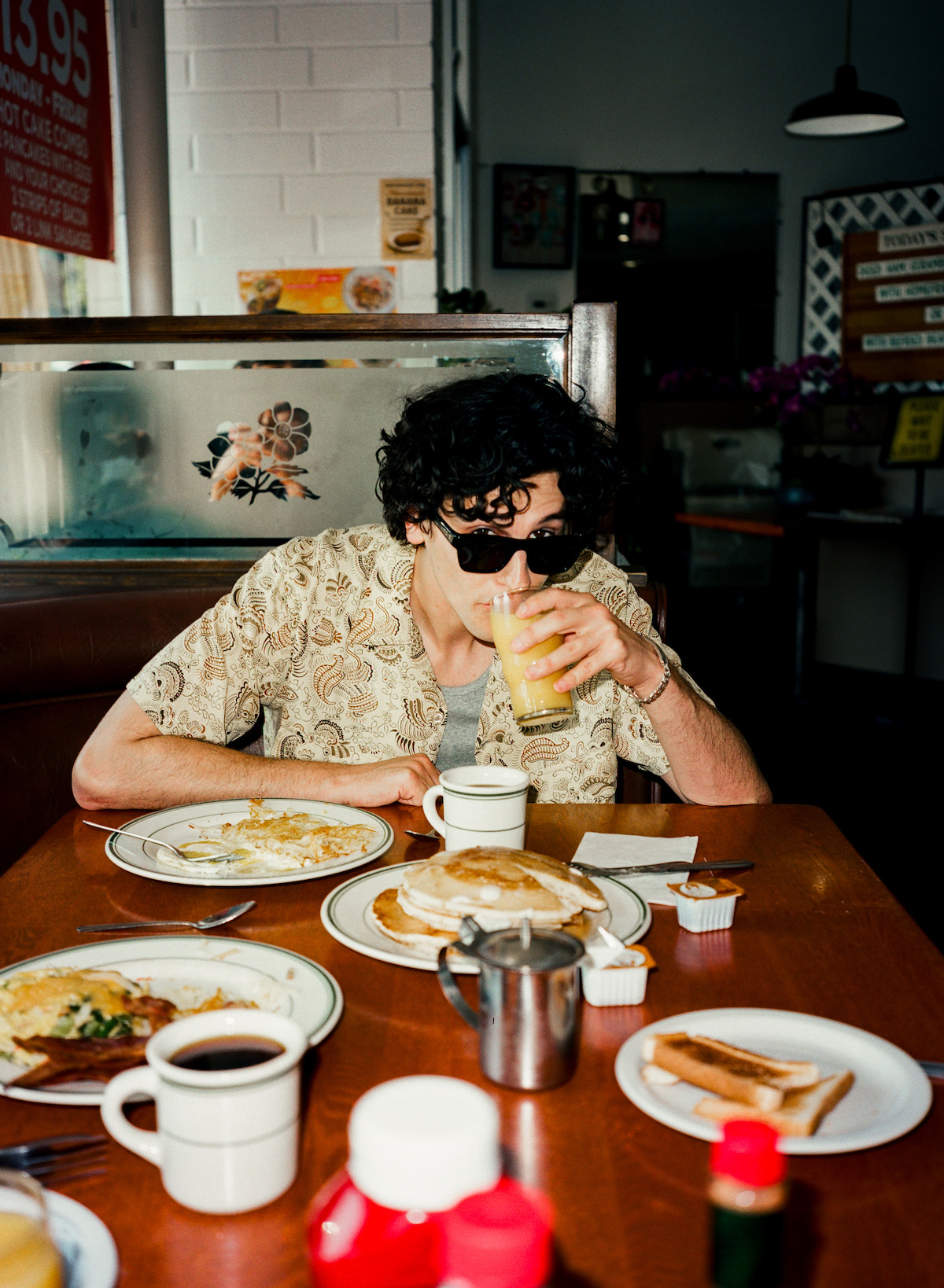 Owen Painter wears sunglasses and drinks orange juiceat a diner table