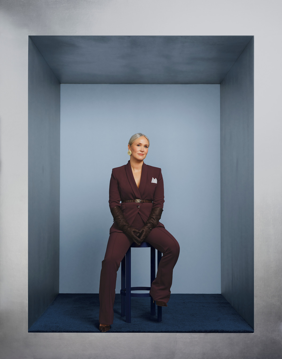 Woman in a burgundy suit and gloves sits on a stool in a minimalist, monochromatic gray-blue box-shaped room, posing confidently with a composed expression in a modern, studio setting.