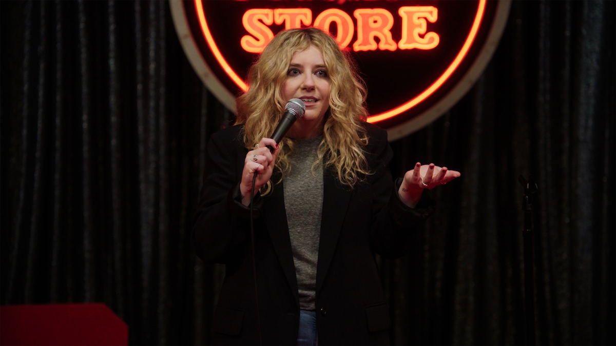 A woman with curly blonde hair holds a microphone while performing stand-up comedy on stage, with a neon sign that says "STORE" in the background and dark curtains behind her.