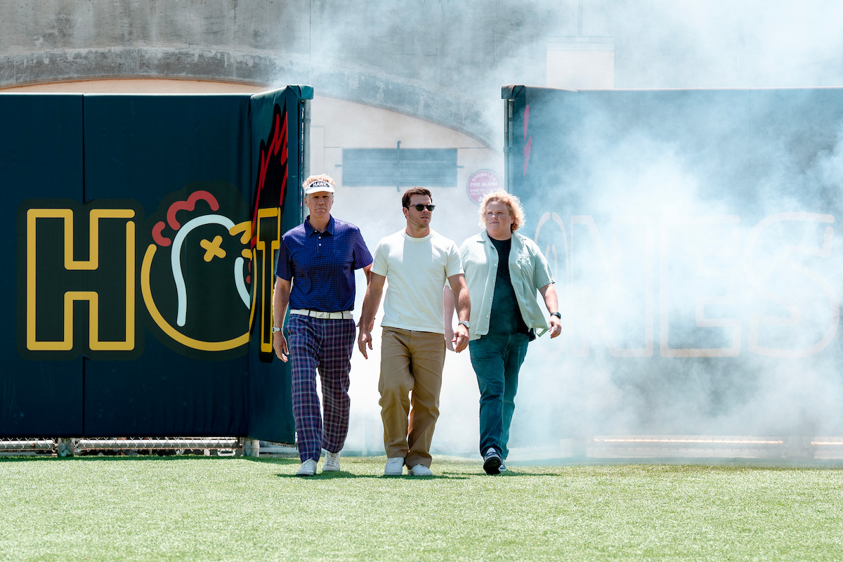 Three men walk confidently out of stadium tunnel onto grass field, one wearing sunglasses, smoke or fog surrounds them, colorful sign with chicken graphic and large yellow letters in background, midday outdoor setting.