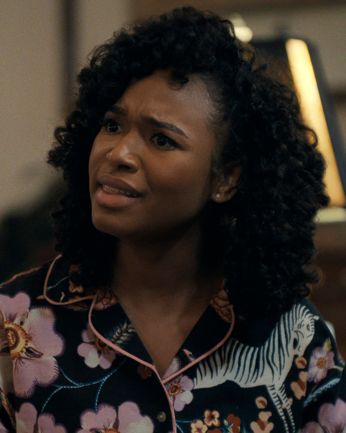 Young woman with curly hair wearing a floral and zebra print shirt, indoors, looking slightly confused or concerned. The background appears to be a warmly lit room with blurred furniture and decor.