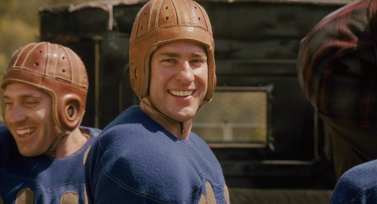 Smiling football players in vintage leather helmets and blue uniforms stand outdoors, likely on a field, with an old vehicle in the background, suggesting a historical or early 20th-century football setting.