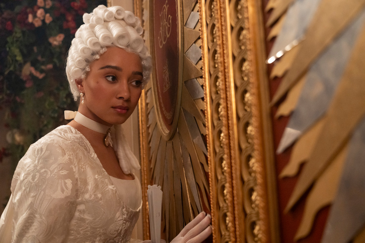 Woman in ornate white period costume and wig stands indoors, closely facing a decorated golden wall, with floral decorations blurred in the background, evoking an 18th-century historical setting.
