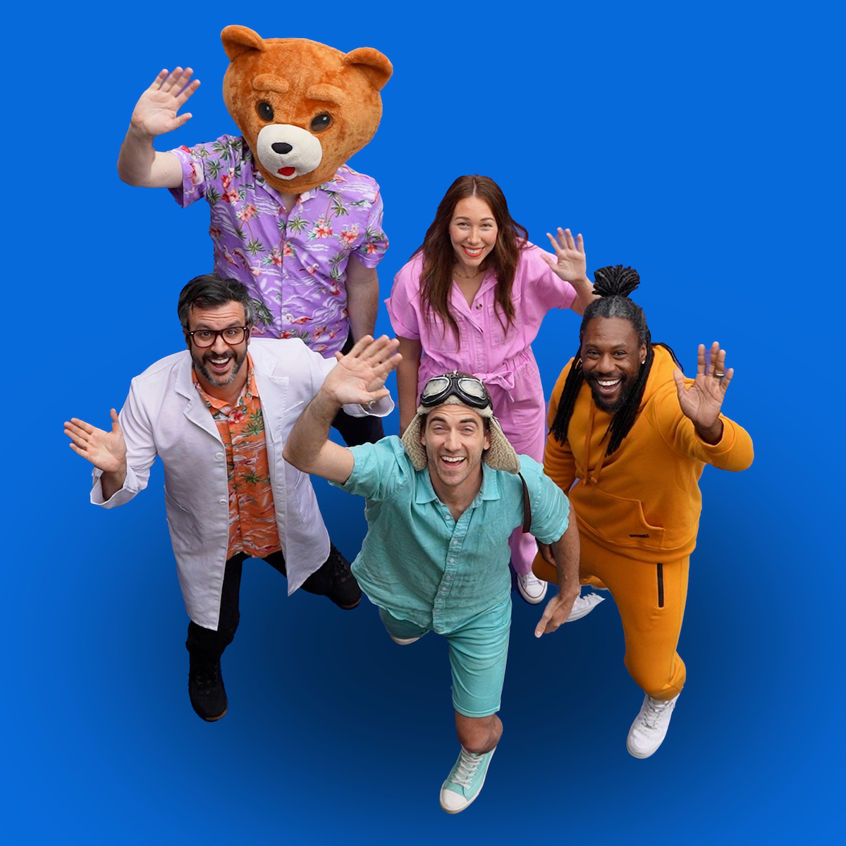 Five adults in colorful costumes, including one in a bear mascot suit, stand on a bright blue background waving and smiling enthusiastically at the camera, suggesting a fun and playful group environment.