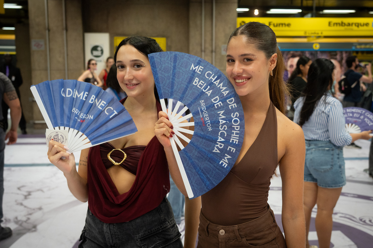 Duas moças sorriem em um local público movimentado, provavelmente uma estação de transporte, enquanto seguram leques decorativos azuis com texto em português. Elas aparecem cercadas por outras pessoas ao fundo.