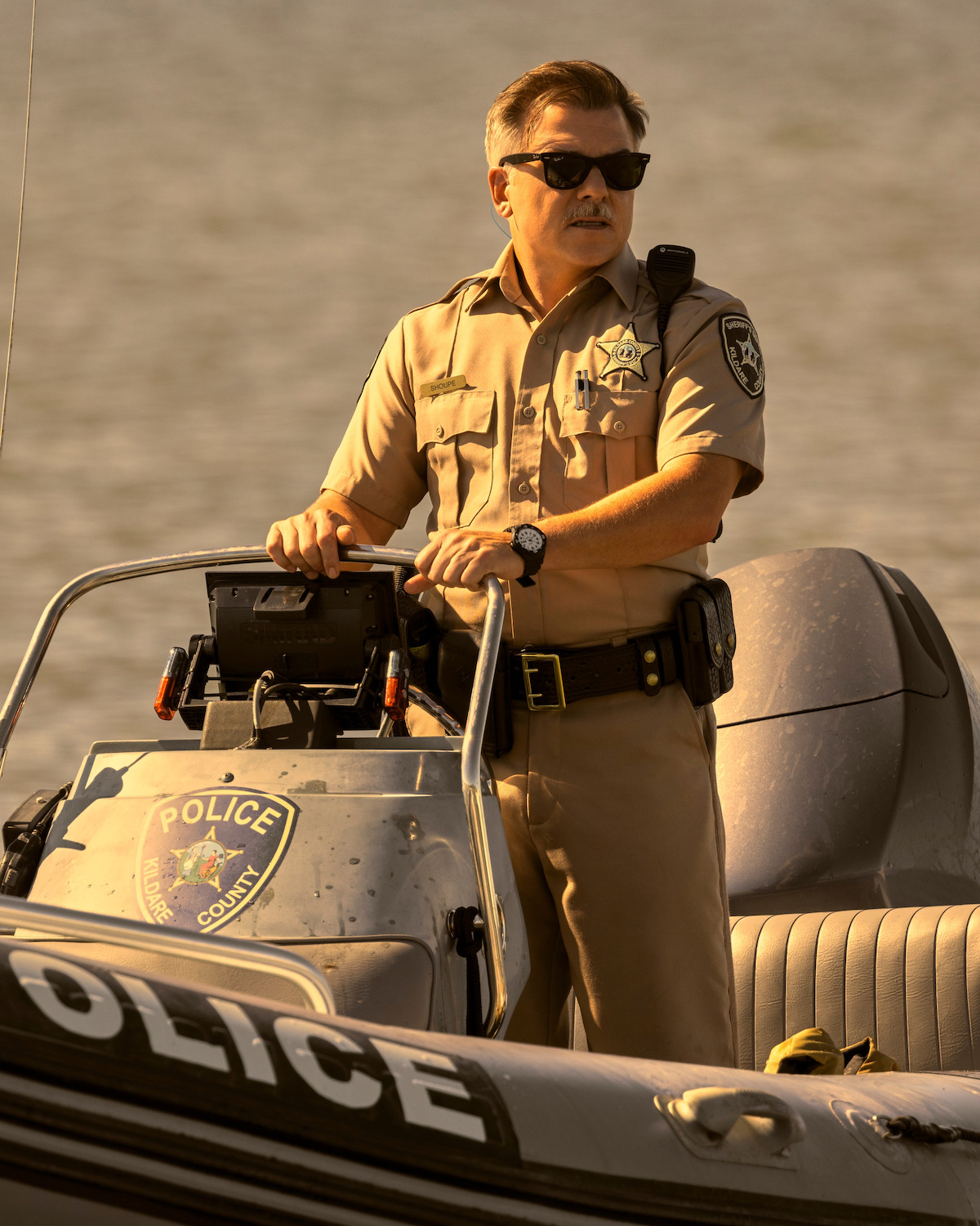 Cullen Moss as Deputy Shoupe in uniform and sunglasses driving a police boat on a body of water, with sunlight creating a warm tone on the scene. The boat has police insignia and enforcement equipment visible.
