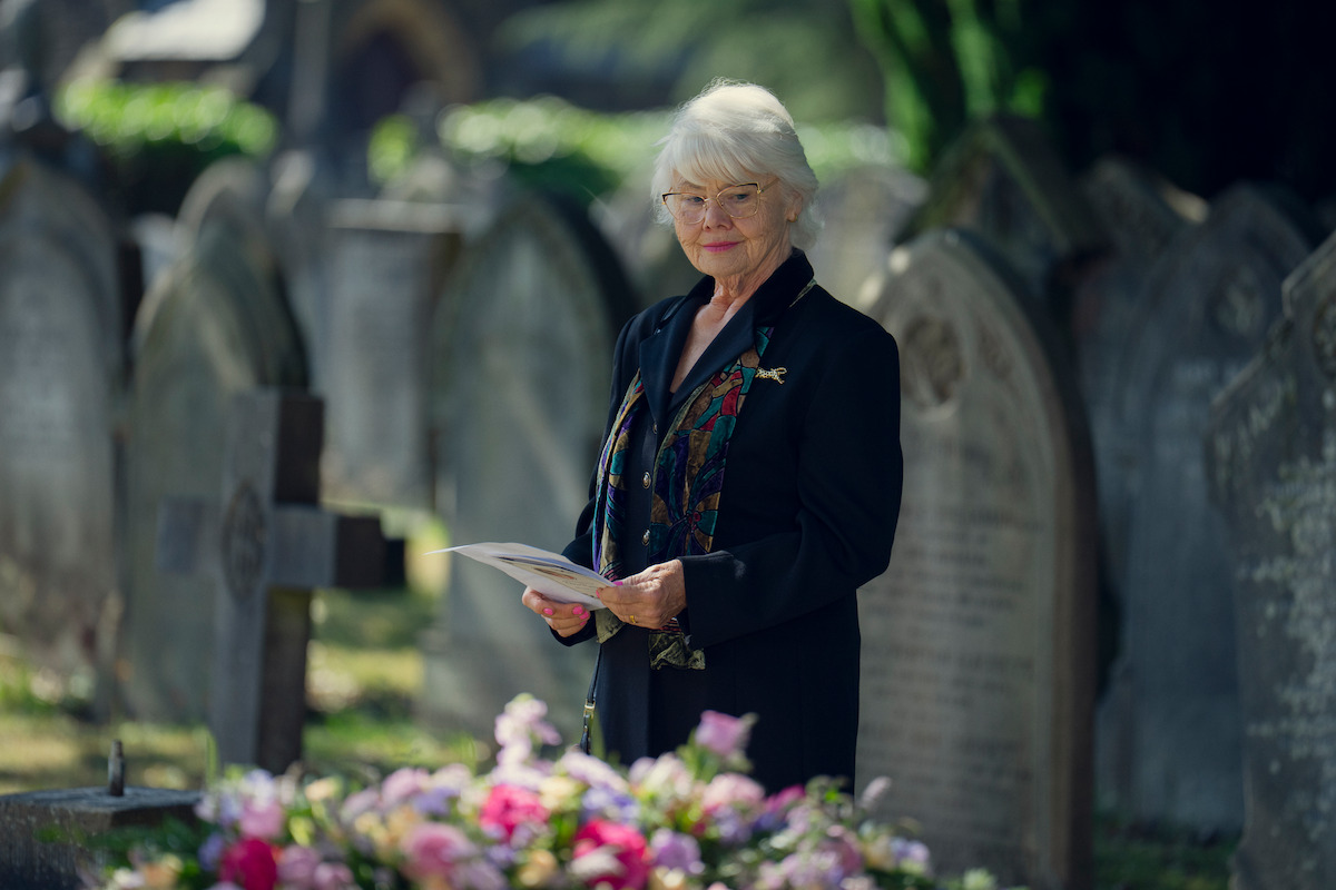 Una anciana vestida de negro sosteniendo papeles se encuentra en un cementerio cerca de lápidas y una tumba adornada con flores coloridas, rodeada de vegetación y luz solar.