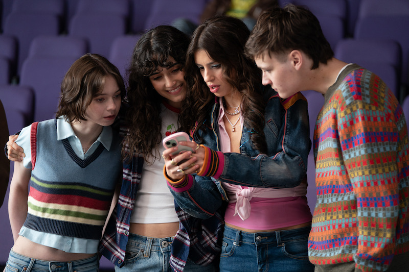 Four teenagers in casual clothes standing together indoors, closely looking at a smartphone, appearing engaged and curious, with rows of purple seats in the background, suggesting a school or auditorium setting.