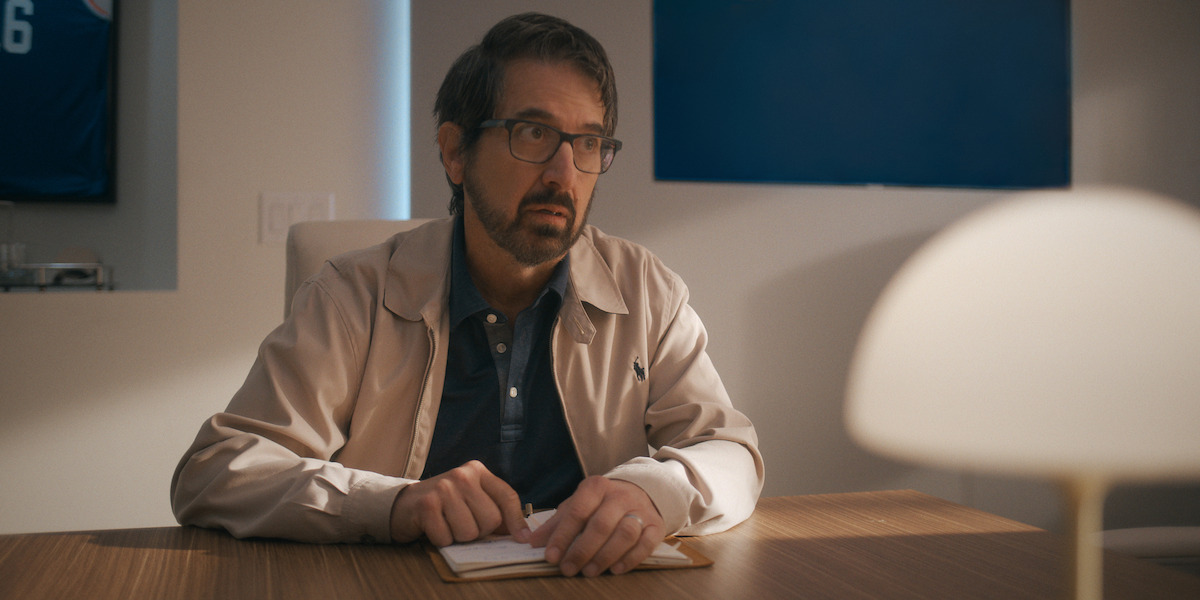Man with glasses and beard sits at a wooden desk holding a notepad, in a modern office with blue accents and soft lamp light.