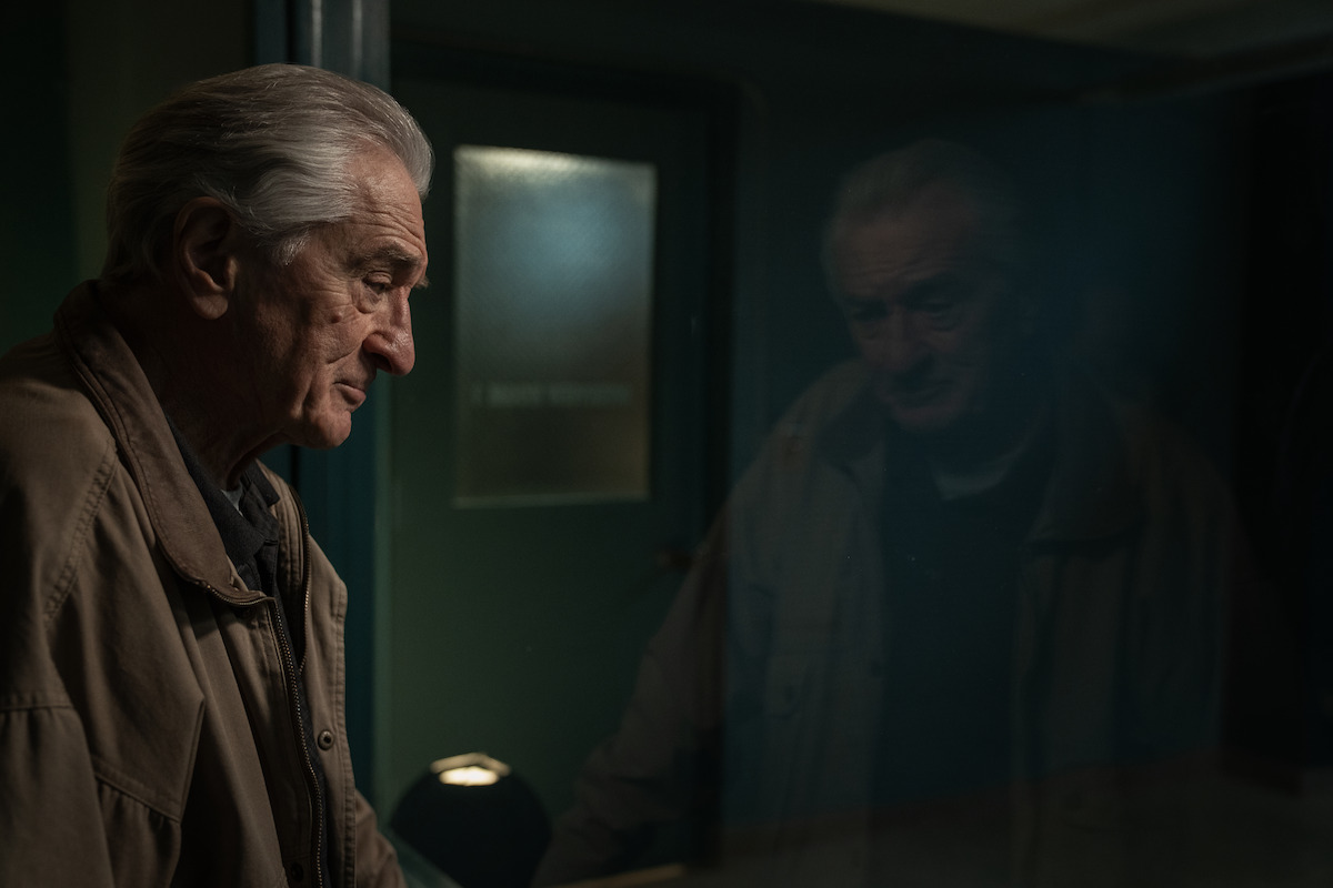Elderly man in a tan coat stands in a dimly lit room, his reflection visible in a glass pane. The setting appears somber and introspective, with dark tones and a closed door in the background.