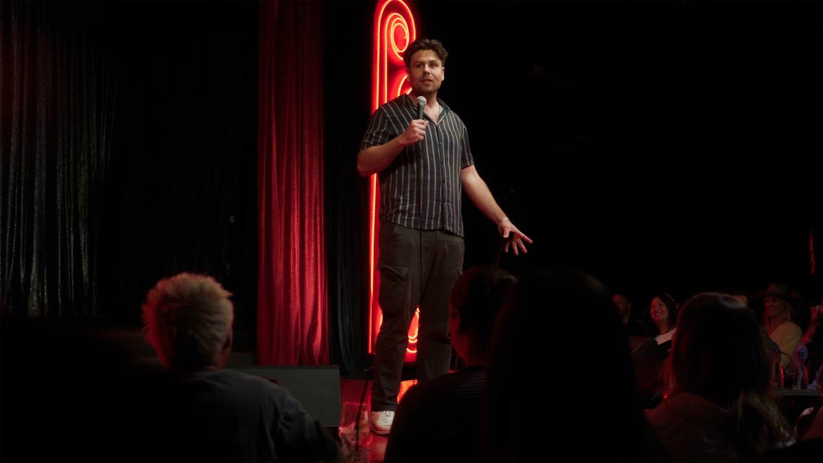 Comedian performing on stage with microphone in hand, neon red light and curtains in background, audience seated in dimly lit comedy club setting.
