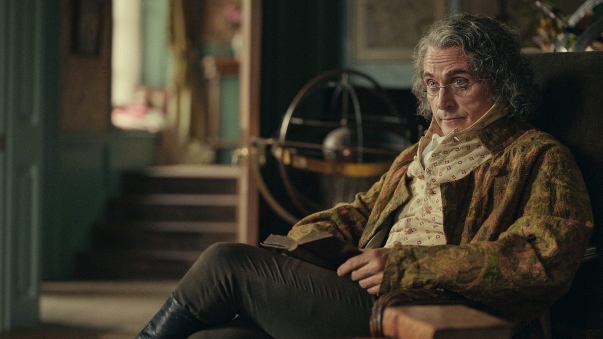 Older man with curly gray hair and glasses sits in an ornate, vintage room holding a book, surrounded by antique furnishings, with an armillary sphere and staircase in the background, evoking a historical or scholarly atmosphere.