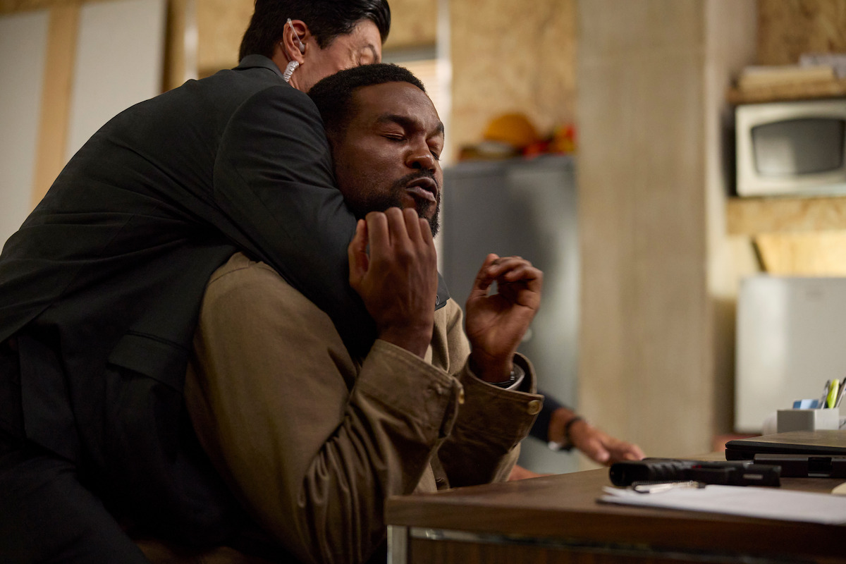 Two men struggle in an office; one man in a suit restrains another in a chokehold near a cluttered desk with a gun and papers, with shelves and office equipment in the background.