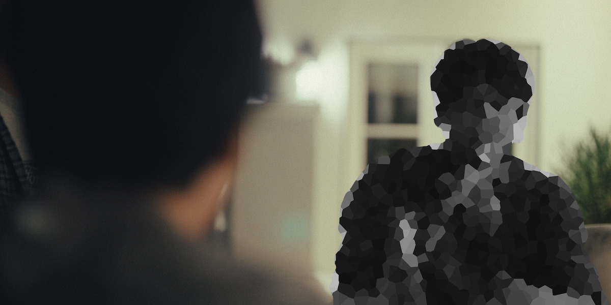 Two people interact indoors; one in the foreground is out of focus while the other, seated in a well-lit living room, is pixelated and indistinguishable. There are plants and windows in the background.