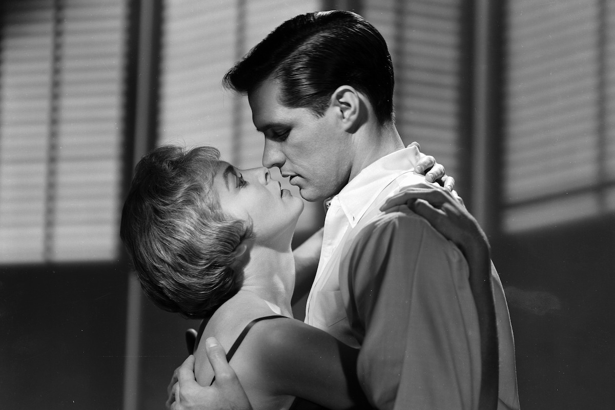 A man and woman embrace and prepare to kiss in a dramatic black and white scene, standing indoors with window blinds in the background.
