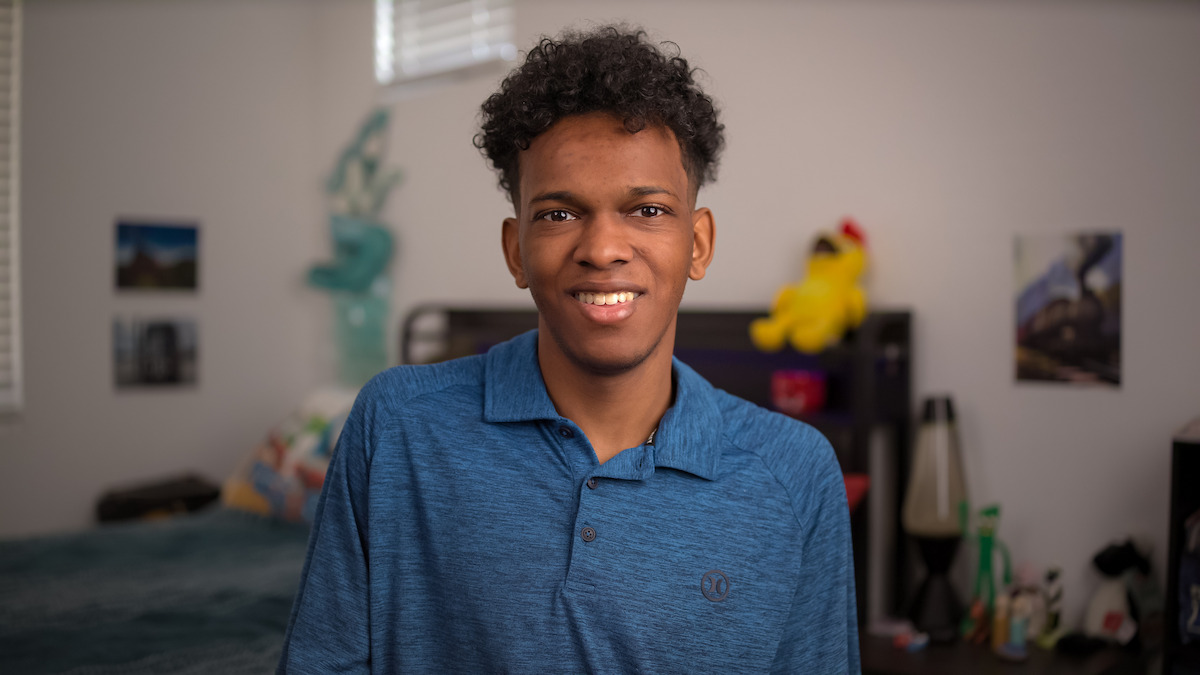Young man smiling in a bedroom with a blue polo shirt, decorated with stuffed toys, posters, bed, lamps, and window in the background, creating a casual and cozy setting.