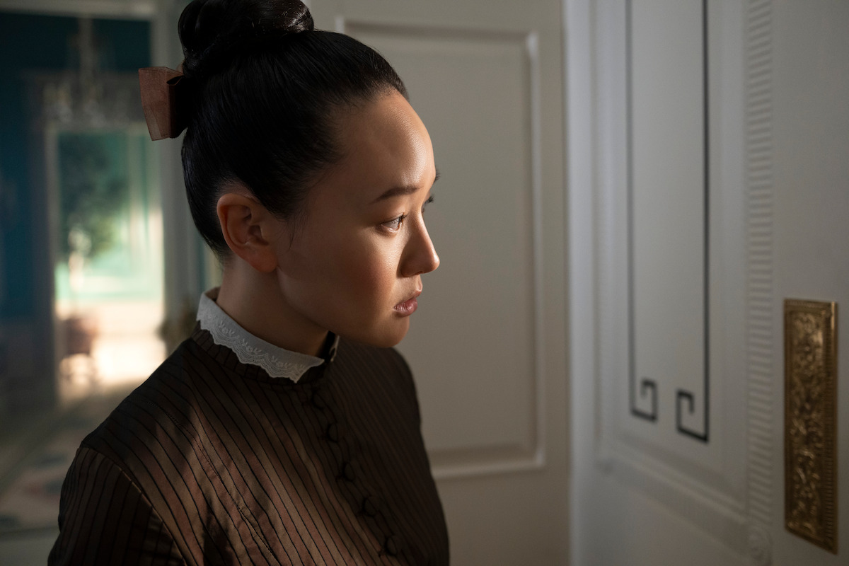 Woman in period clothing stands indoors by a white paneled door, looking intently toward an ornate light switch in a softly lit, elegant room.