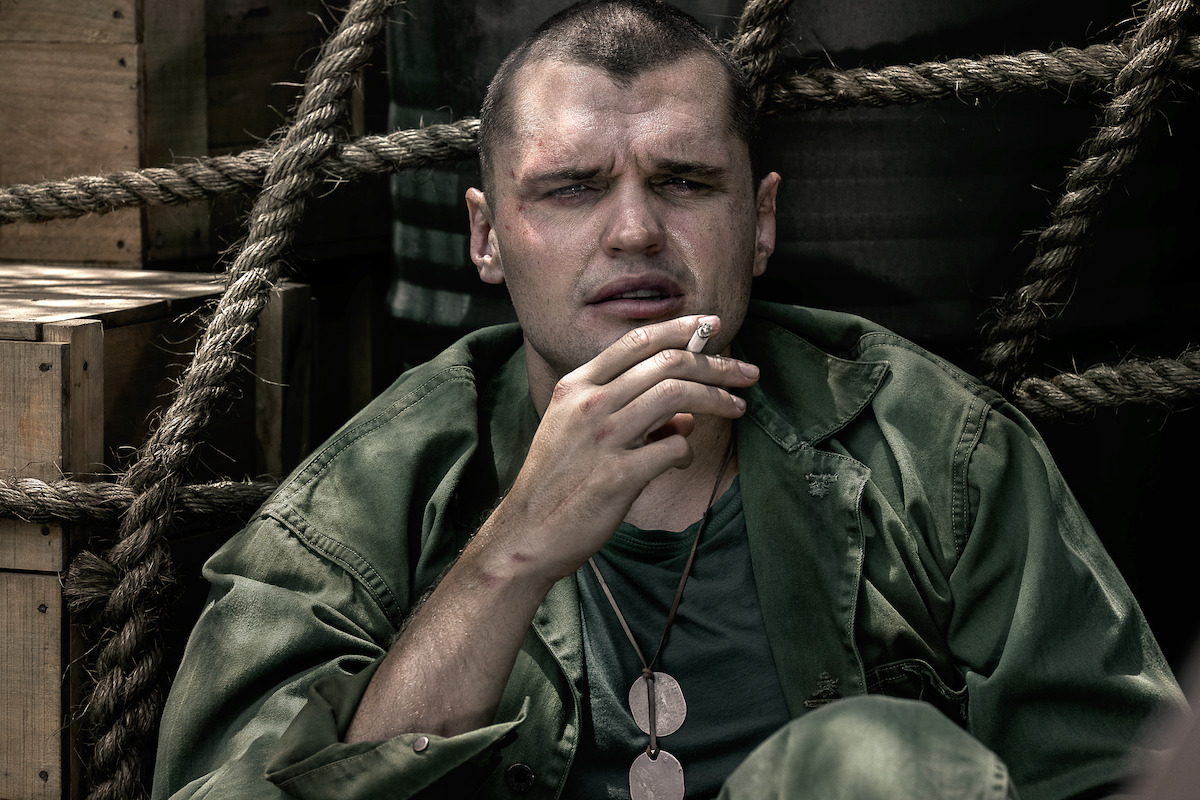 Ray Nicholson as Tony Butkovich in green fatigues sits against wooden crates and ropes, holding a cigarette. He wears dog tags and has a serious expression, suggesting a tense or contemplative moment in a military setting.