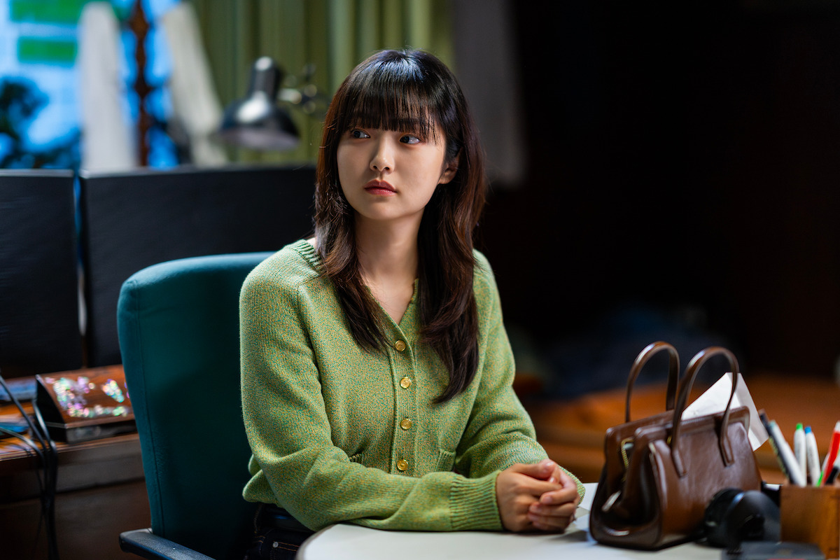 Young woman with long dark hair in a green sweater sits at a desk in a softly lit office, looking to the side. A brown handbag and office supplies are on the desk. The atmosphere feels calm and contemplative.