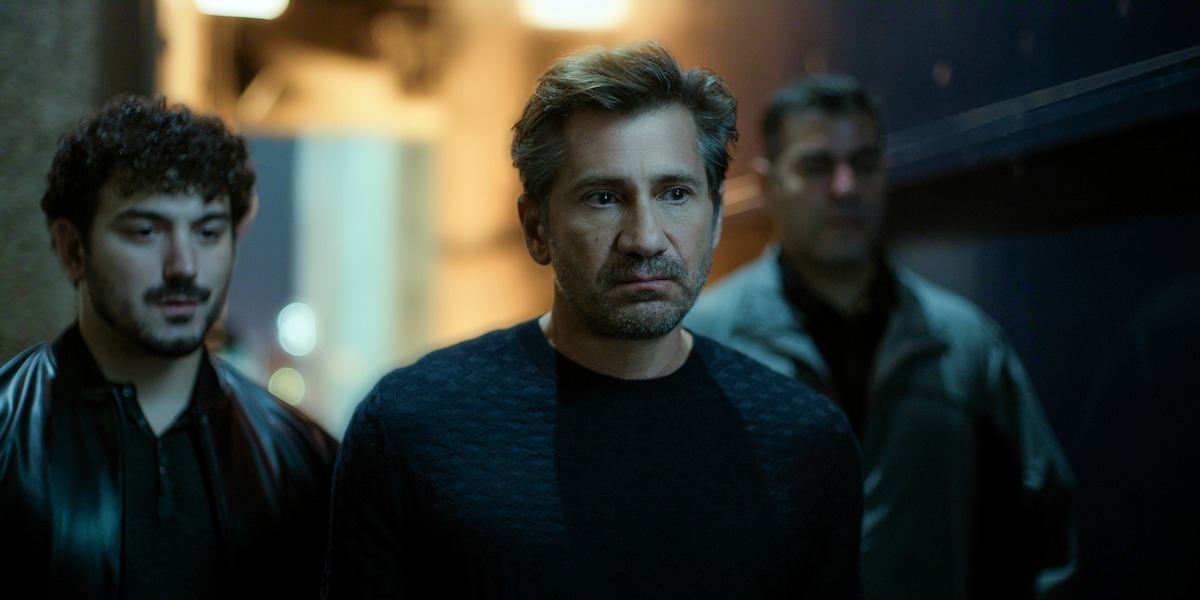 Three men with serious expressions stand in a dimly lit hallway, wearing dark clothing. The background is out of focus, adding a tense and dramatic atmosphere to the scene.