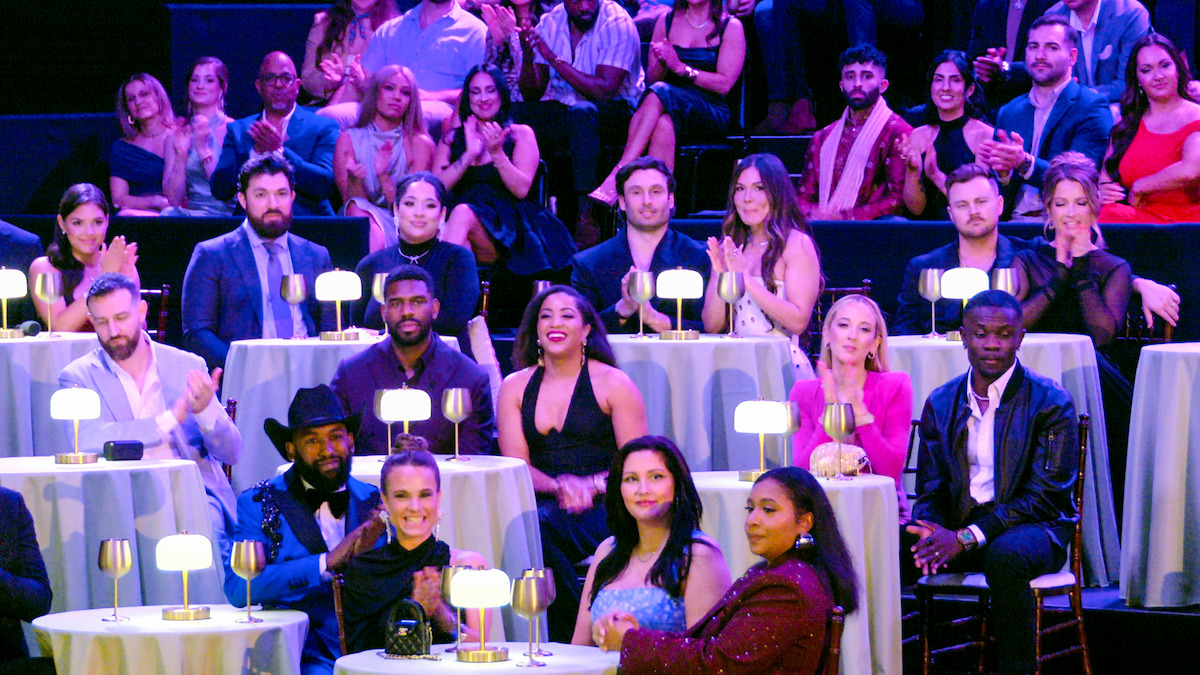 A diverse group of people dressed formally, seated at round tables with glowing lamps in a dimly lit event venue, watching and applauding a performance or presentation on stage.