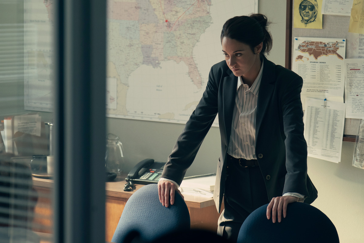 A woman in a suit leans on a conference table in an office, appearing serious and focused. The background features a large map, bulletin board with notes, a desk phone, and documents, suggesting an investigative or professional setting.