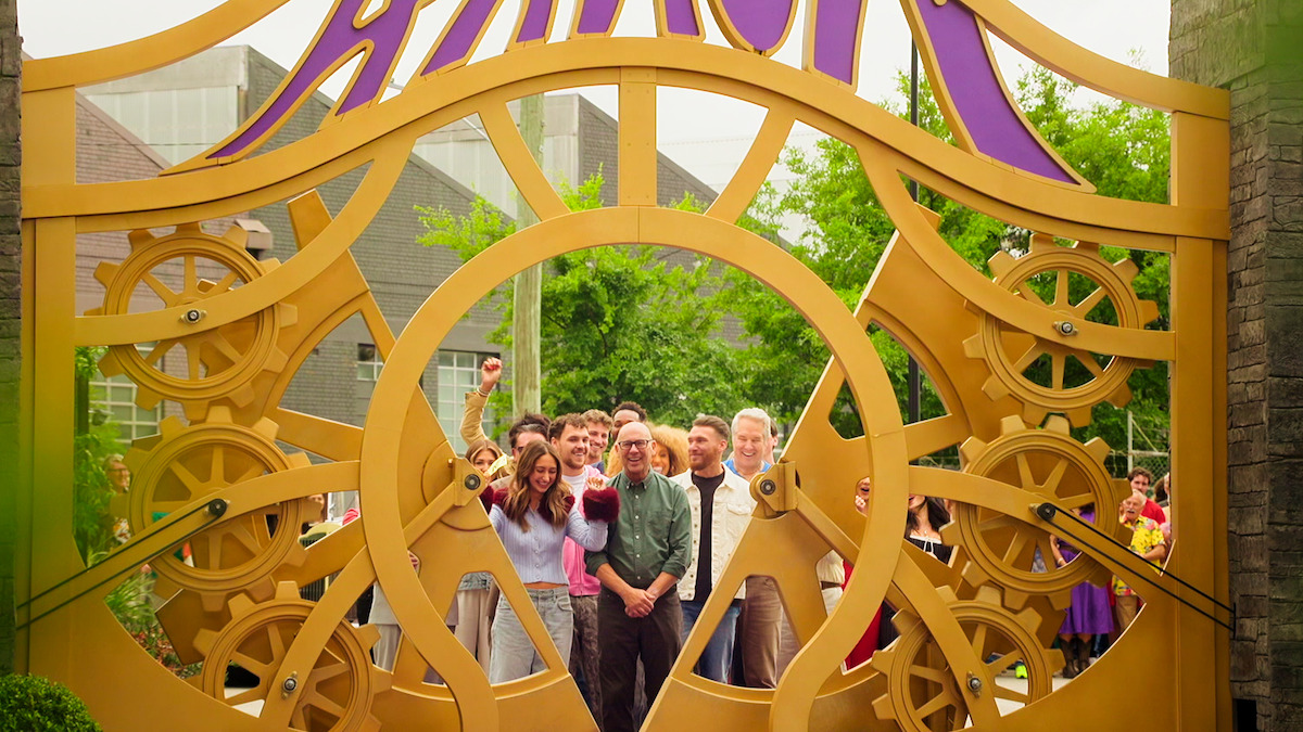 A group of people smiling and posing together behind a large decorative gate with gears and the word "Amuck" above them, set outside with trees and buildings in the background.