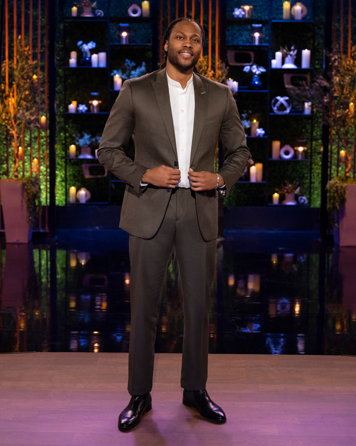Man in a brown suit, standing on a polished stage with decorative plants and shelves filled with candles and ornaments in the background, giving a formal and elegant atmosphere.