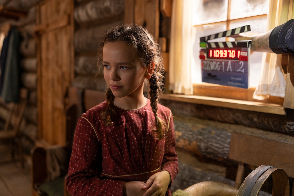 Young girl with braided hair wearing a red dress stands in a rustic wooden cabin set, with a film clapperboard visible by the window, indicating a movie or TV production in progress. Warm lighting, vintage setting.