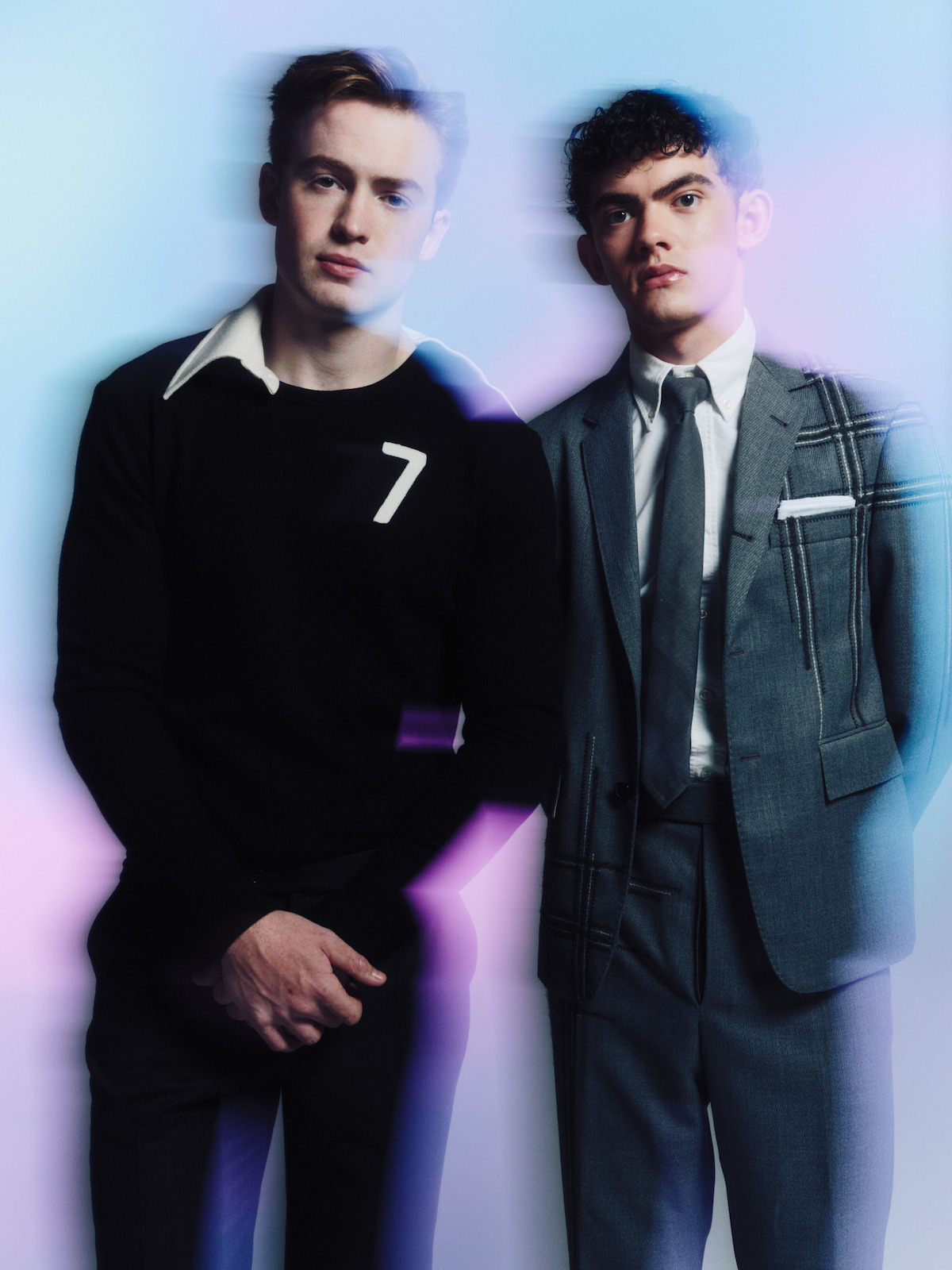 Two young men stand side by side against a softly lit, pastel gradient background. One wears a black shirt with a number 7, the other a stylish gray suit and tie. The image has a dreamy, slightly blurred effect.