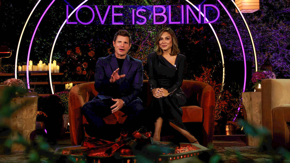 Two hosts sit on a stylish set with glowing neon "Love Is Blind" sign, surrounded by candles and warm colors, creating an inviting, romantic, and welcoming atmosphere for a television show.