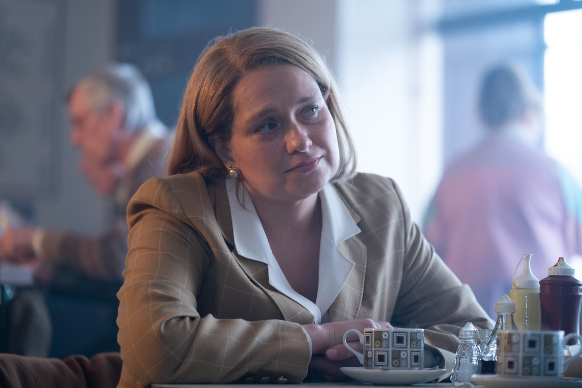 Woman in a tan blazer sits at a café table with patterned coffee cups, salt, pepper, and condiment bottles. The background shows other people seated, giving a relaxed, social atmosphere with soft natural lighting.